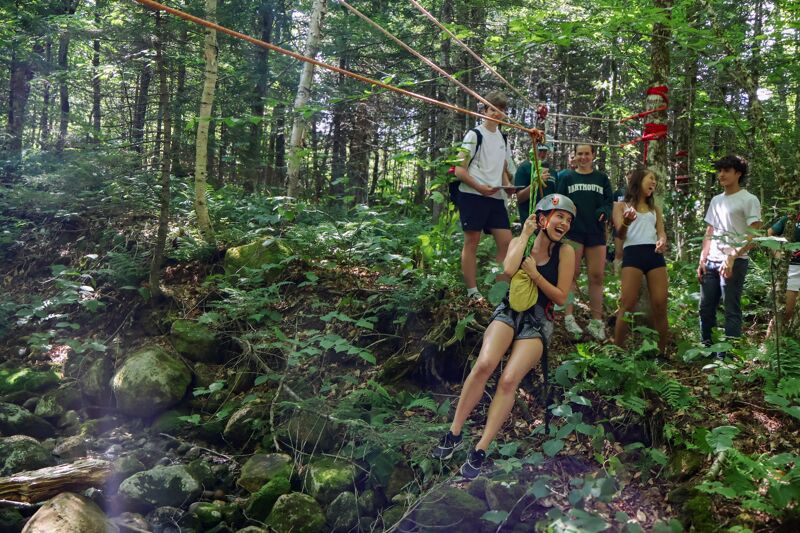 The image shows a group of people enjoying a zip line adventure in a forest. A woman is in the middle of the zip line, while others are watching. The scene is filled with lush greenery and trees, suggesting an outdoor recreational activity.
